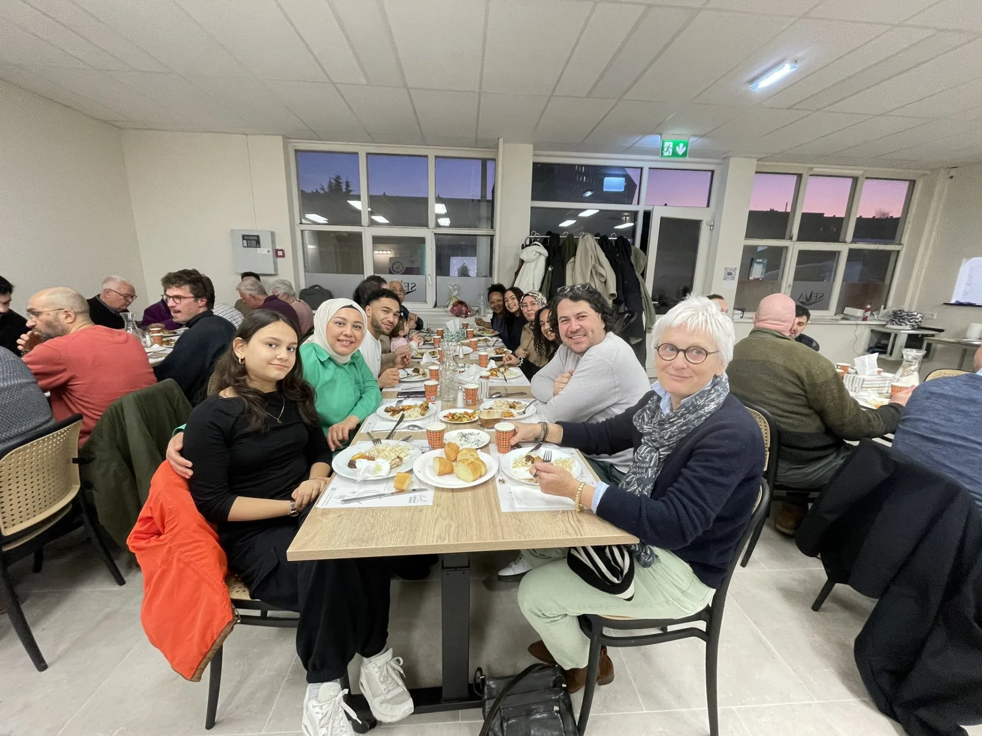 Gasten aan lange tafels tijdens de eerste iftar in de vernieuwde zaal van SEVA